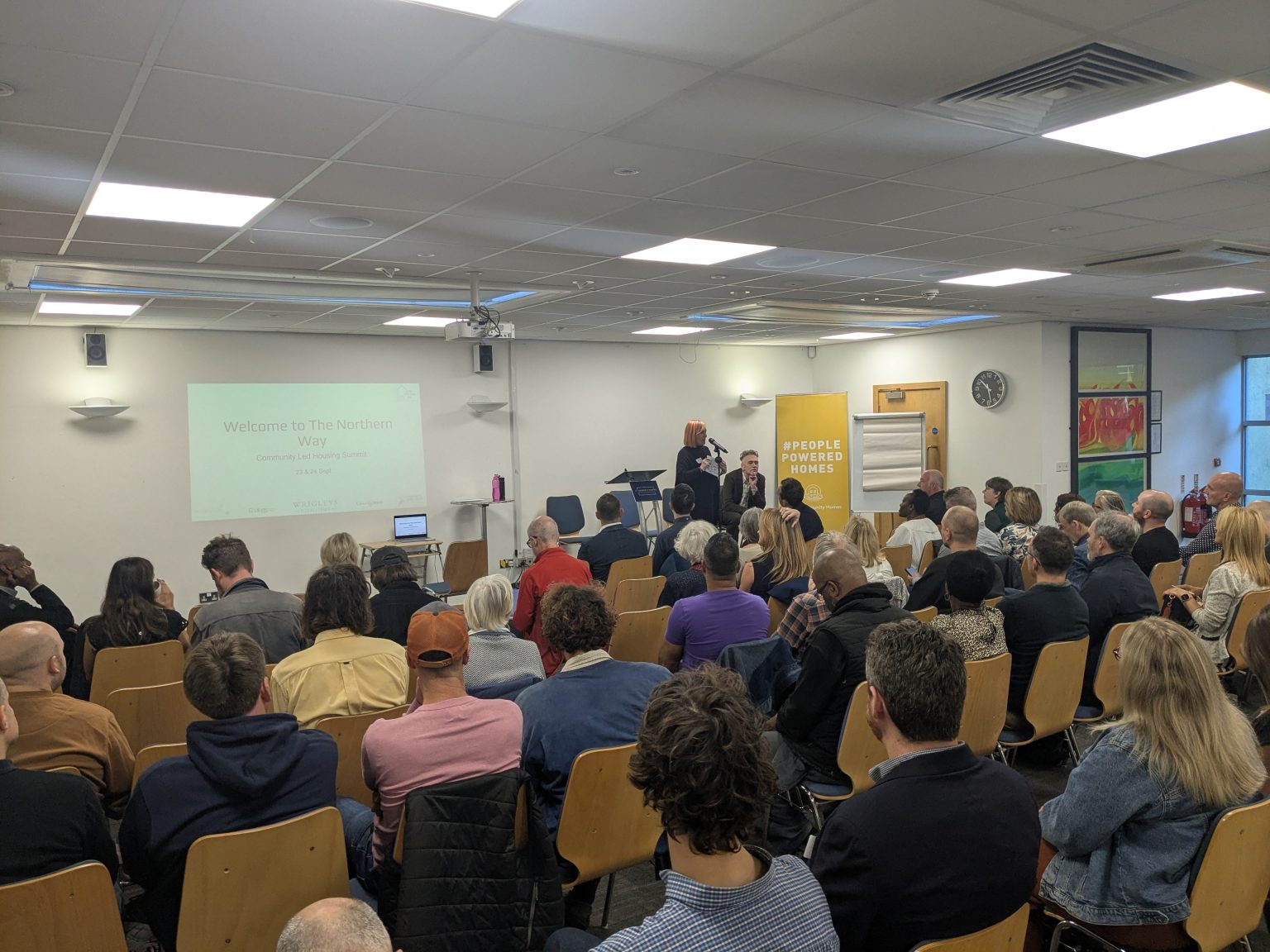 An image of the back of the audience at the Northern Way Summit. They are sat inside a room looking at a presentation that says Welcome to the Northern Way Summit. Peg Alexander is hosting the session. 
